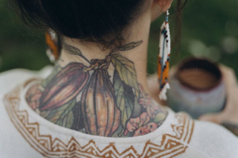 Angelina Katharina Petersen von hinten Fotografiert, man sieht ihren tätowierten Nacken, dort sind zwei Kakaoschoten zu sehen, sie hält vor sich eine Tasse zeremoniellen Rohkakao