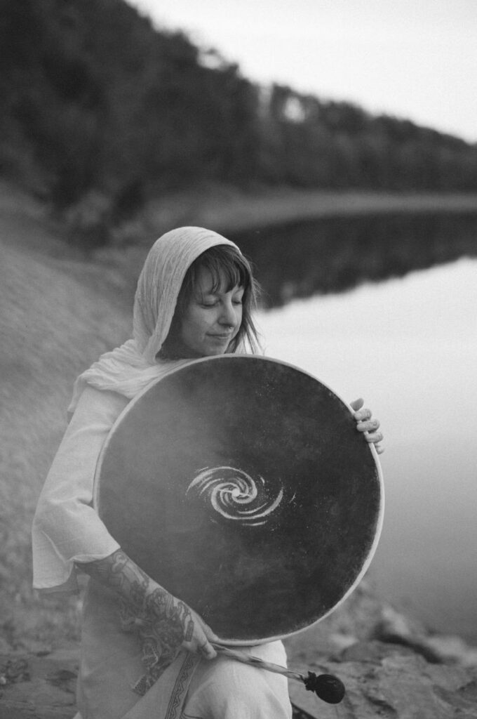 Schwarz-weiß-Porträt von Angelina Katharina Petersen in Leipzig, im weißen Leinenkleid und Kopftuch mit geschlossenen Augen liebevoll ihre Trommel mit Spiralgalaxie haltend, im Hintergrund Wasser und Natur
