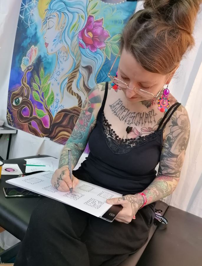Angelina Katharina Petersen auf einer Tattoo-Convention in Leipzig, sitzend mit Brille auf einer Liege beim Zeichnen von Motiven, im Hintergrund das große Florina Venus Banner
