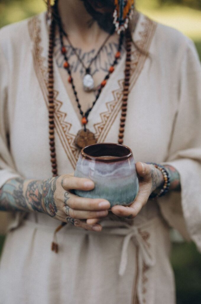 Angelina Katharina Petersen in Leipzig hält im weißen Leinenkleid eine Keramiktasse mit zeremoniellem Kakao, ihre Mala- und Ayahuasca-Kette sichtbar