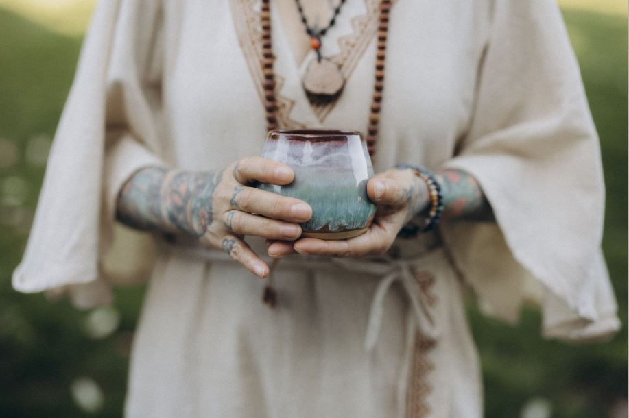 Angelina Katharina Petersen in Leipzig hält im weißen Leinenkleid eine Keramiktasse mit zeremoniellem Kakao, ihre Mala- und Ayahuasca-Kette sichtbar