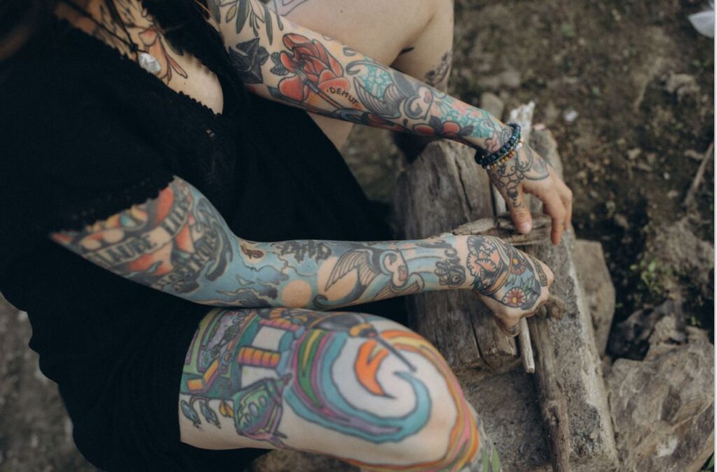 Angelina Katharina Petersen in Leipzig, sitzend im schwarzen Kleid vor Holzscheiten, mit tätowierten Beinen, Armen und Blume am Dekolleté